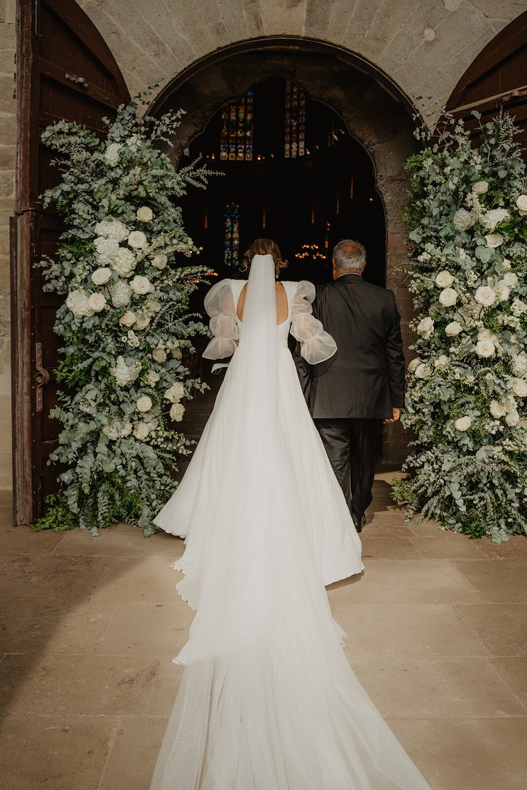 Novia entrando en una iglesia histórica catalana decorada con rosas blancas y eucalipto